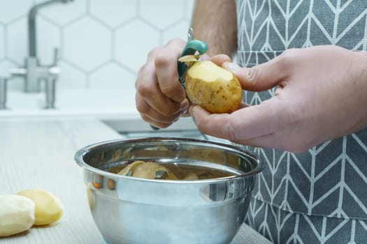 Cropped photo of man wearing grey apron peeling potato with green peeler over metal bowl full of peelings, standing at grey wooden table in kitchen. Recycling, composting, organic waste management.