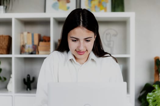 Brunette woman working with laptop at home or office. Girl is reading computer screen and smiling. Restrained joyful face. Blurred background with furniture. Person working from home concept