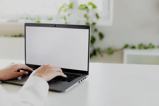 Woman hands typing on the laptop with empty blank white screen mockup. Computer is laying on the white table. Blurred background. Person working from home concept. Copy space area for text