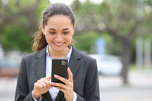 Front view portrait of a happy businesswoman using mobile phone walking in the street