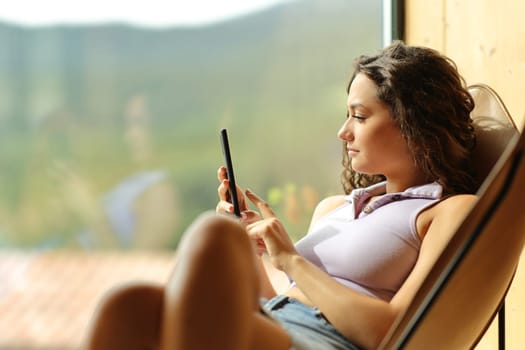 Woman using phone sitting on a chair at home beside a window