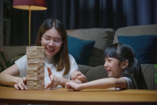 Smiling woman help teach child play build constructor of wooden blocks, Asian young mother playing game in wood block with her little daughter in home living room at night time before going to bed
