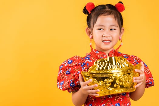 Chinese New Year. Happy Asian Chinese little girl smile wearing red cheongsam holding big gold ingot, Portrait children in traditional dress hold golden bar, studio short isolated on yellow background