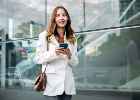 Asian businesswoman working with mobile phone standing against street front building near office, Portrait successful business woman smiling holding smartphone use application chat online in morning