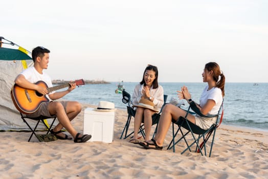 Happy friend have fun playing guitar and clap in camp they smiling together in holiday on sand beach near camping tent vacation time at sunset, Young Asian group woman and man in summer travel outdoor