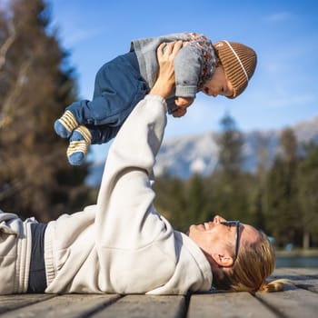 Happy family. Young mother playing with her baby boy infant oudoors on sunny autumn day. Portrait of mom and little son on wooden platform by lake. Positive human emotions, feelings, joy