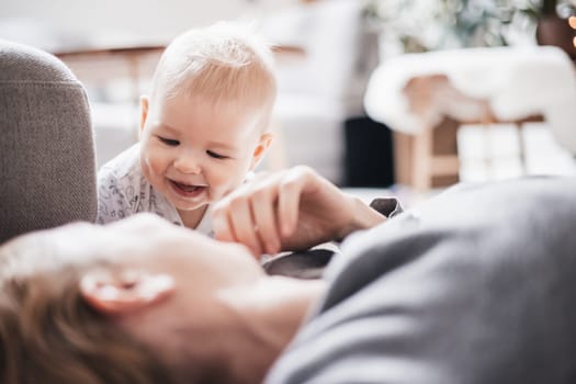 Happy family moments. Mother lying comfortably on children's mat playing with her baby boy watching and suppervising his first steps. Positive human emotions, feelings, joy