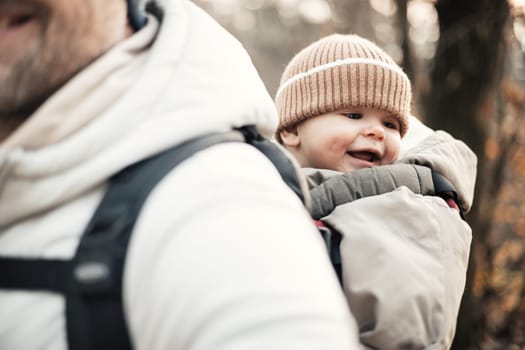 Sporty father carrying his infant son wearing winter jumpsuit and cap in backpack carrier hiking in autumn forest