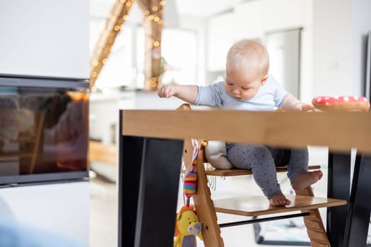 Happy infant sitting at dining table and playing with his toy in traditional scandinavian designer wooden high chair in modern bright atic home. Cute baby playing with toys.