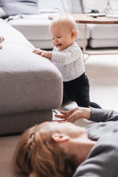 Happy family moments. Mother lying comfortably on children's mat playing with her baby boy watching and suppervising his first steps. Positive human emotions, feelings, joy