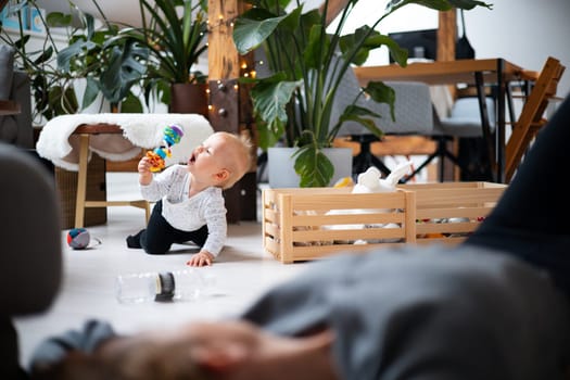Happy family moments. Mother lying comfortably on children's mat watching and suppervising her baby boy playinghis in living room. Positive human emotions, feelings, joy.