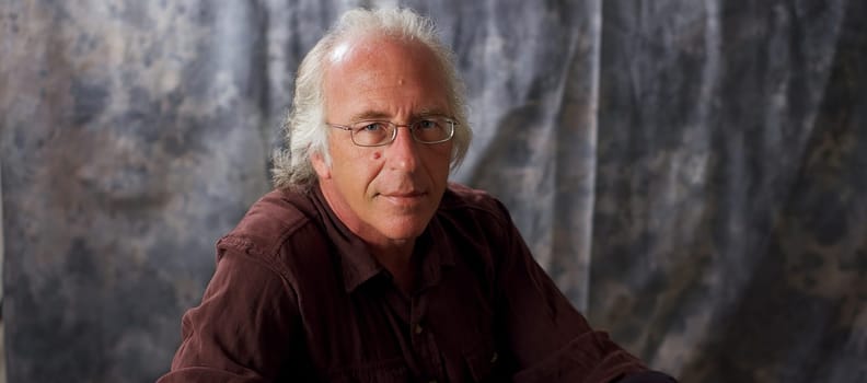 An older blue eyed man with glasses and white hair wearing a brown shirt against mottled gray background.