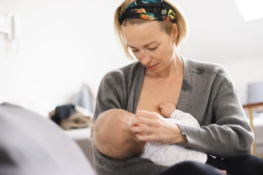 Young woman breastfeeding her infant baby boy casualy sitting on child's playing mat on living room floor at home