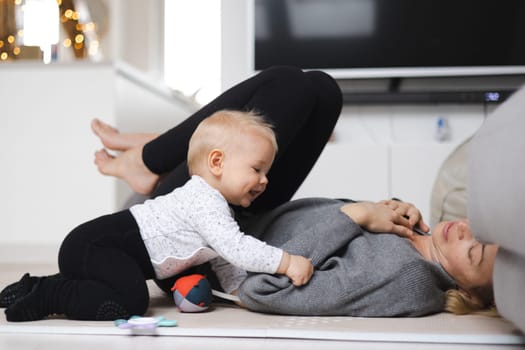 Happy family moments. Mother lying comfortably on children's mat playing with her baby boy watching and suppervising his first steps. Positive human emotions, feelings, joy