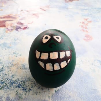 Easter egg dyed green with a painted laughing face. Cool grimace with eyes and big white teeth. Scary mug for Halloween. Emoticon for Easter. Light abstract background