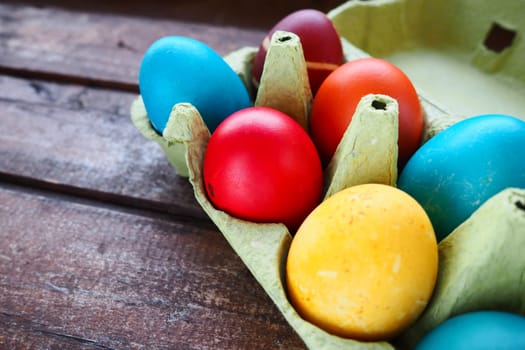 Multi-colored eggs painted with gouache and onion peel in a cardboard box on a wooden table background. Ten boiled eggs. Postcard poster for Easter. Easter holiday. Red, blue, yellow, brown eggs.