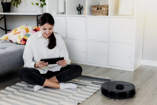 Young Caucasian woman using tablet, chatting, playing while robot vacuum cleaner is cleaning floor in the living room. Pretty girl is configuring device using mobile application over wifi network