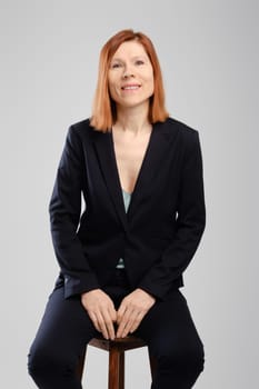 Stylish middle aged woman in pantsuit sitting in studio over grey background