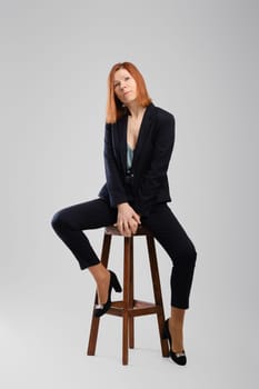 Stylish middle aged woman in pantsuit posing in studio sitting on chair with legs spread over grey background