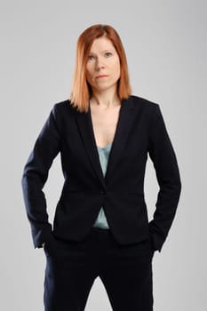 Stylish middle aged woman in pantsuit posing in studio with hands in pockets over grey background