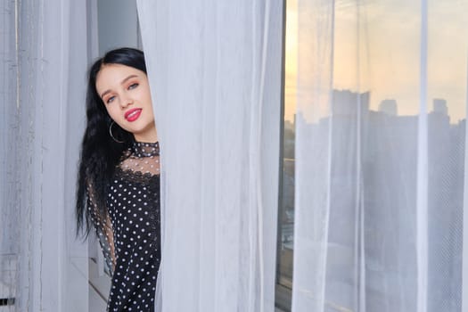 Cute young woman woman peeking out of the curtains