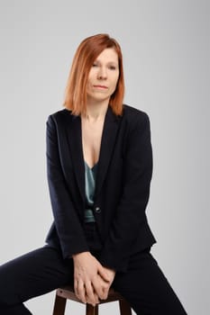 Stylish middle aged woman in pantsuit sitting in studio over grey background