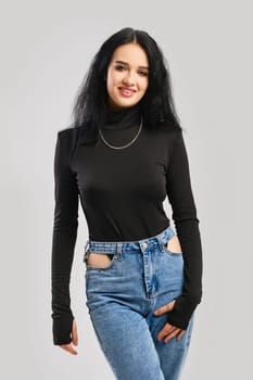 Young happy woman posing in studio in black bodysuit with long sleeves and oversized jean over grey background