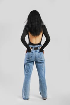 Back view of barefoot woman with hands in pockets in studio in black bodysuit with long sleeves and oversized jean over grey background
