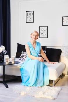 Retired woman in long blue dress sits on couch at home in sunny day