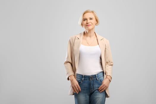 Confident middle age woman standing with arms in the pockets looking at camera at gray background