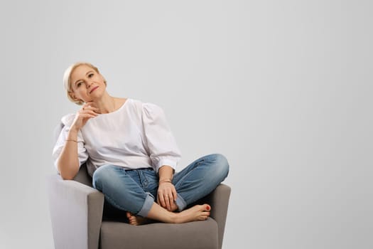 Relaxed senior woman sitting in an armchair crossing legs in studio