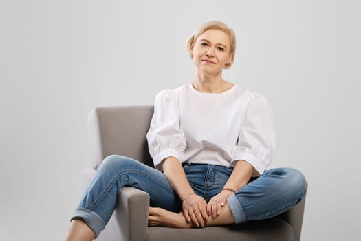 Positive mature woman sitting in an armchair in studio, looking at camera