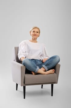 Barefoot senior woman sitting in an armchair crossing legs in studio