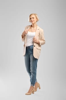 Full length portrait of senior woman in white tank top, beige jacket and boyfriend jeans over grey background