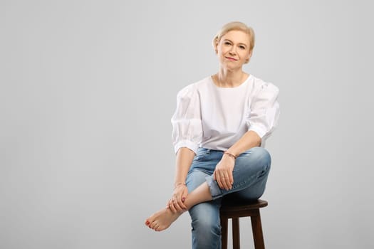 Barefoot senior woman sitting on tall chair crossing leg in studio