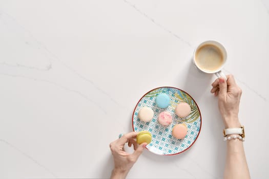 Top view of colorful cake macaroons on a plate and female hand with cup of coffee