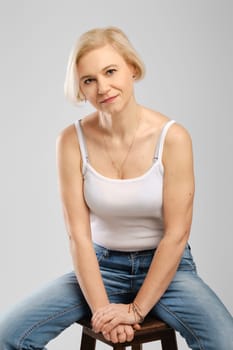 Barefoot senior woman in jeans and tank top sitting on tall chair in studio
