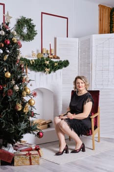 Middle age woman sitting in cosy armchair near christmas tree and fireplace