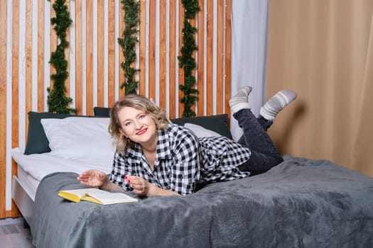 Cheerful middle age woman in casual wear lays on bed and reading book