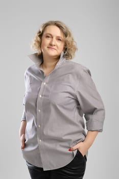 Casual portrait of senior woman in grey shirt and trousers with hands in pockets over grey studio background
