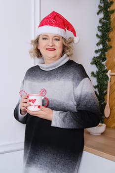 Middle age woman in christmas cap standing in small kitchen and holds a cup with hot beverage in hand