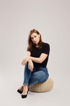 Pensive woman in casual black t shirt and jeans sits on the studio floor over grey background