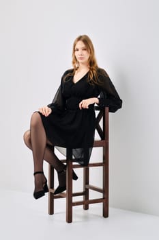 Full length portrait of cute young woman in black tulle dress sitting in profile on tall chair in white studio