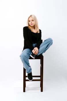 Teen girl in tank top, jacket and jeans posing on chair in white studio