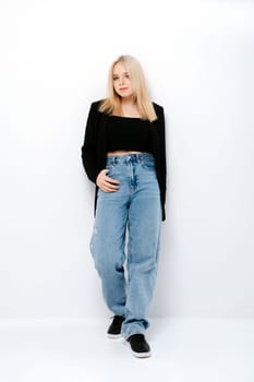 Blonde girl in tank black top, jacket and jeans posing over white wall