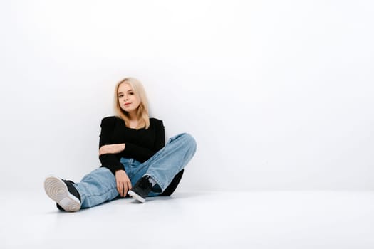 Cute girl in tank top, jacket and jeans posing sitting on the floor in white studio