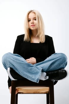 Tricky girl in tank top, jacket and jeans sits on chair with folded legs over white background