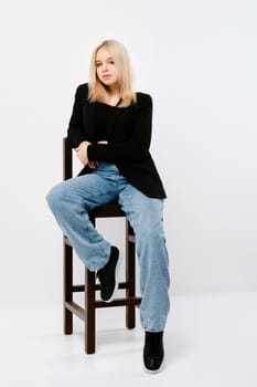 Teenager girl in tank top, jacket and jeans sits on chair in white studio