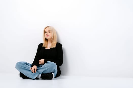 Pensive girl in tank top, jacket and jeans posing sitting on the floor with crossed legs in studio