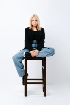 Teen in tank top, jacket and jeans sits on tall chair in white studio